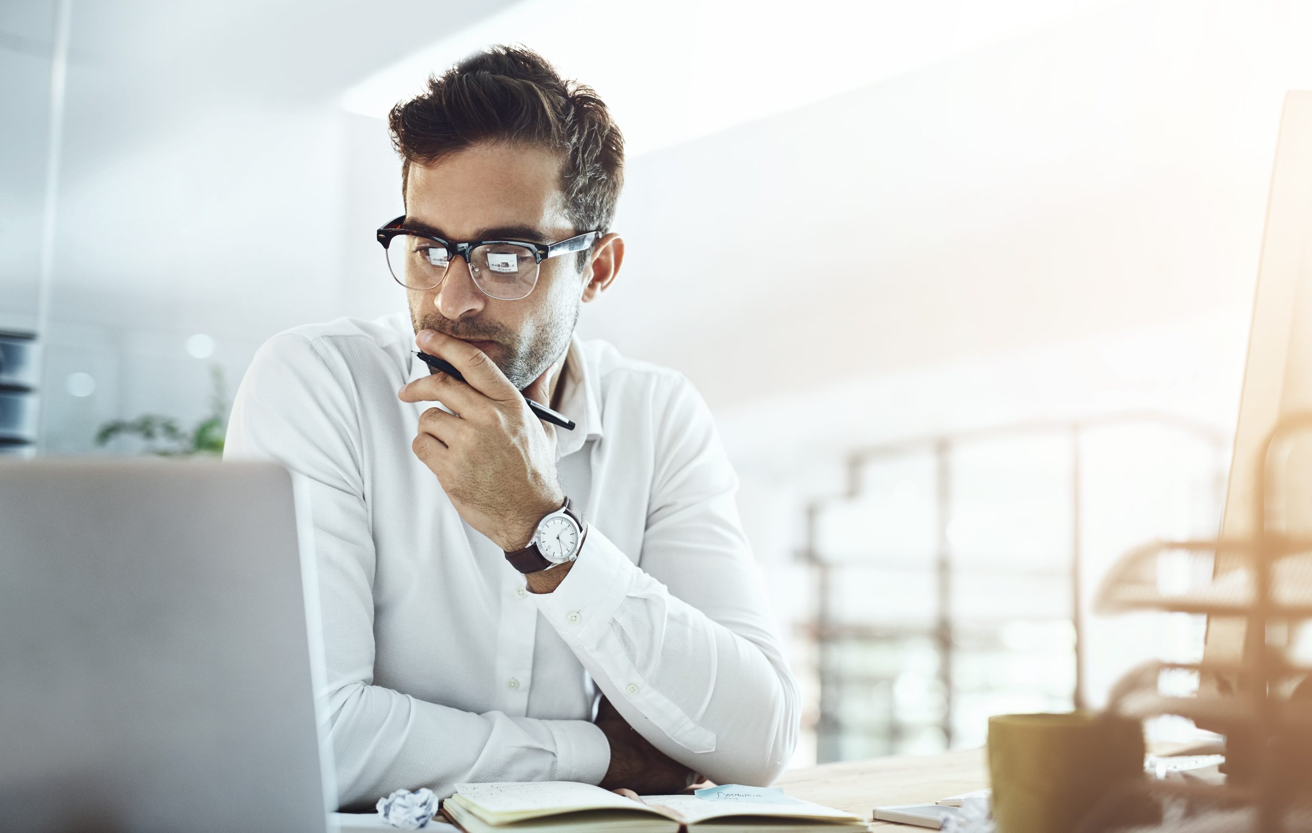 A man wearing glasses and a white shirt is sitting at a desk with his hand on his chin, intently analyzing debt recovery strategies on a laptop screen. An open notebook and a cup are on the desk, surrounded by the softly lit ambiance of the room.