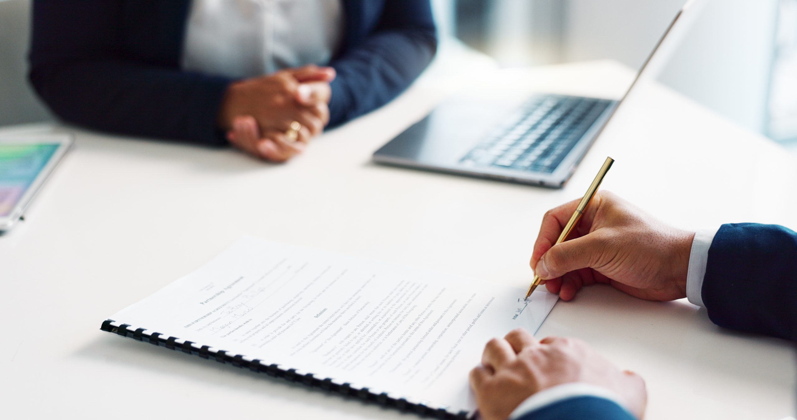 Close-up of a person signing a document at a table, initiating the collections process, while another sits across from them. A laptop is open on the table, and both individuals are dressed in business attire.