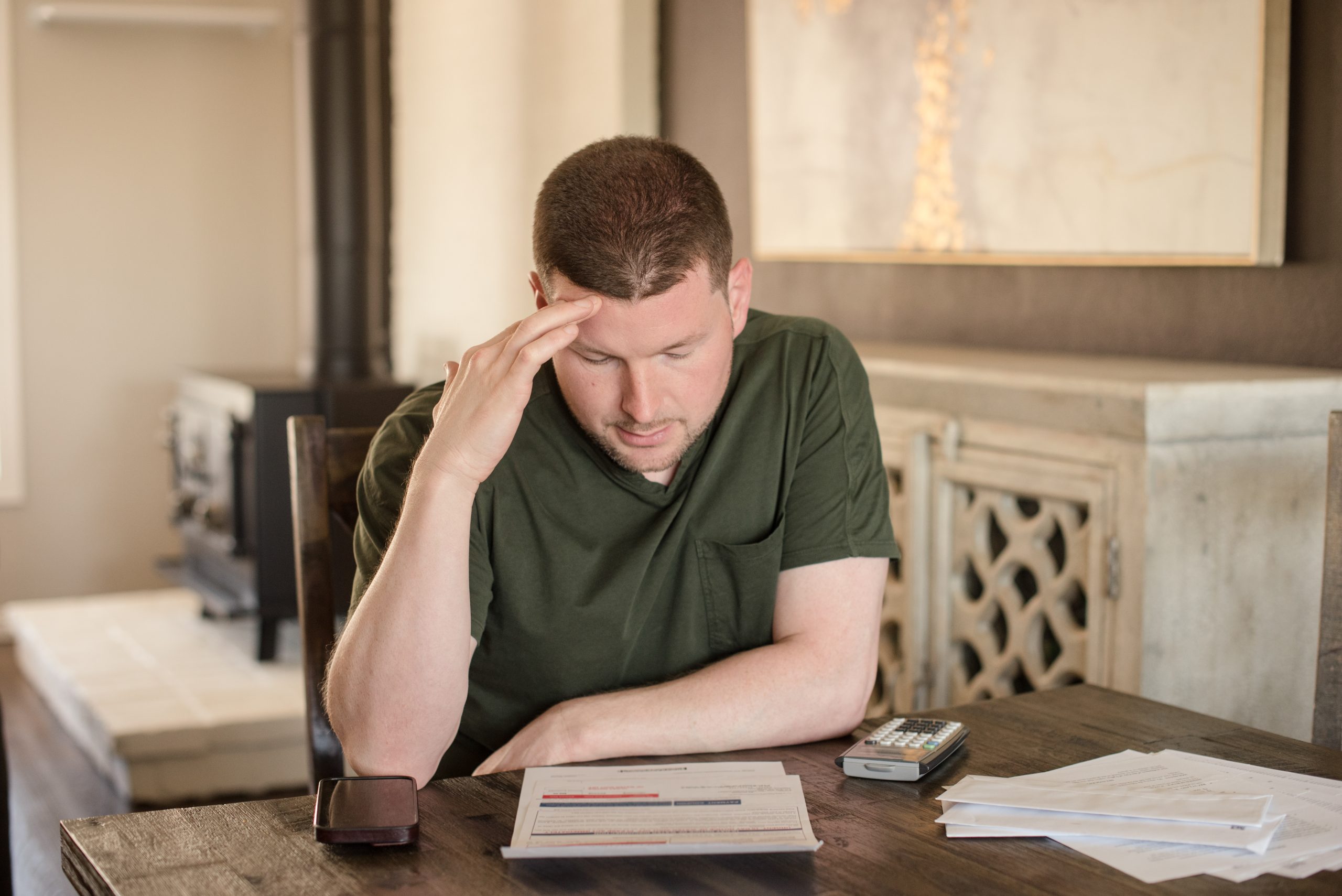 A man in a green shirt sits at a table, looking stressed as he reviews documents related to the collections process. A phone and calculator are on the table beside him. The background features a cabinet and a painting on the wall.